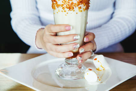 Caucasian female hands with stylish french manicure holds glass of caramel milk shake coctail covered with chocolate, whipping cream, waffles and straw. Cropped frame.の写真素材
