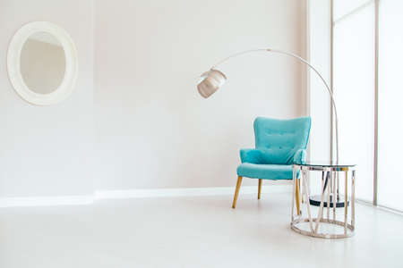 Conceptual light  interior of living room with blue armchair, metallic standing lamp,  glass round coffee table , oval mirror on white wall and large window. Concept of minimalizm.の写真素材