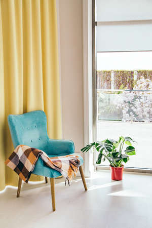 Conceptual light  interior of living room with blue armchair and brown blanket, monstera plant in red vase on white floor, large window and yellow curtain.の写真素材
