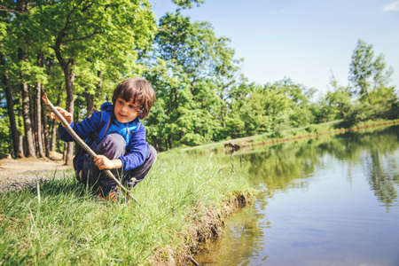Little kid play with stick looks like rod and fishing on the lake in the forest on a sunny day.