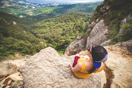 Young woman sitts on  the top of a rock and looks at green valley under.