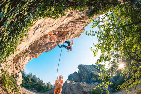 Athletic man climbs a rock, 7c category route in the forest with rope, against sunset and the other...