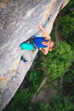 Athletic beautiful blonde hair woman climbs a rock in the forest with rope. Sport climbing, lead....