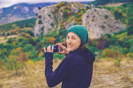 Smiling young traveller woman taking outdoor photo of autumn mountain landscape by her smart phone.の写真素材