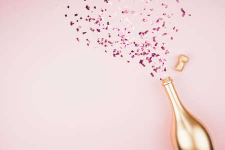 Golden colored bottle of champagne    and splashing out confetti on pink pastel trendy background. Festive background for projects.の写真素材