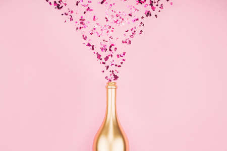 Golden colored bottle of champagne    and splashing out confetti on pink pastel trendy background. Festive background for projects.の写真素材