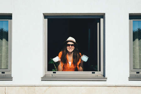 Smiling woman looks out of window and drinks  coffee staying at home. Self isolation concept.の写真素材