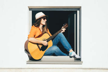 Smiling woman sits in a window and plays the guitar while staying at home. Self isolation concept.の写真素材