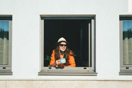 Smiling woman looks out of window and drinks  coffee staying at home. Self isolation concept.の写真素材
