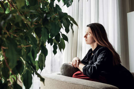 Young self isolated woman sitting at home and looking out of window.の写真素材