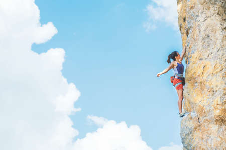 Athletic woman in colorful sportswear climbs a rock with rope on cloud sky background. Sport climbing, lead. Crimea.の写真素材