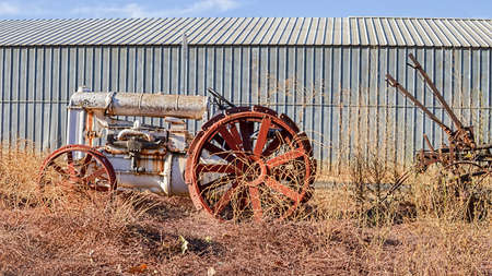 Old white tractor rusting away in old farm equipment graveyard on very dry California grass, against steel wallの写真素材