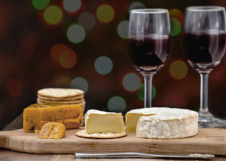 Brie and hot pepper jack cheese on wooden cheese board, accompanied by crackers and two glasses of red wine, against black background, decorated with colorful holiday lightsの写真素材