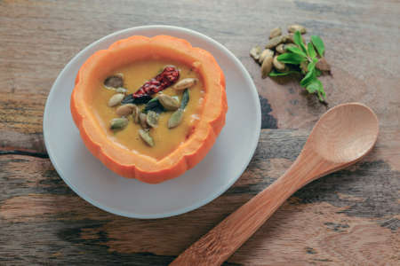 Pumpkin soup served in bowl made of mini-pumpkin, on wooden table, garnished with marjoram, pumpkin seeds and red pepper, ready to be eaten with wooden spoon â?? seasonal food conceptの写真素材