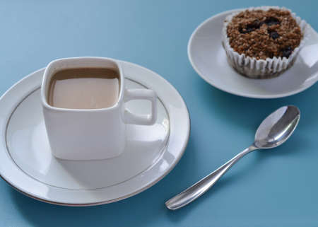 White square cup of coffee, silver spoon and brown fiber muffin set on blue table , viewed from slightly aboveの写真素材
