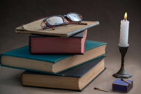 Reading glasses on top of four old books stacked on top of each other, on tan table, against dark background, illuminated with a candlelight - vintage learning at night conceptの写真素材