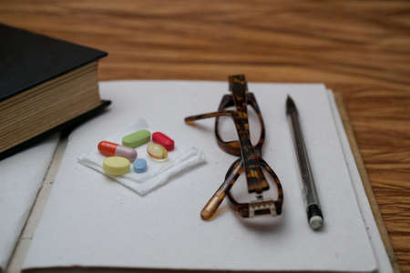 An old book, pair of broken reader glasses on blank journal, a bunch of pills and a pen on wooden table, suggesting an elderly person with memory problems taking a break to take medicationの写真素材