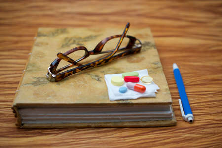 A pair of broken reader glasses on closed journal and a pen on wooden table, suggesting an elderly person taking a break to take medicationの写真素材