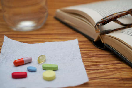 An old hardbound book, pair of broken reader glasses on it, a bunch of pills on white paper towel, on wooden table, suggesting an elderly person taking a break to take medicationの写真素材