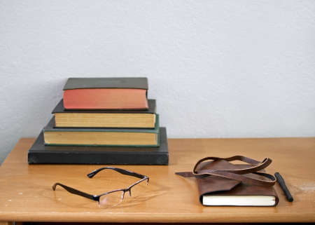 Leather journal, pencil, readers, and hardbound, used books stacked on a weathered, used wooden desk, against white wall - unplugged study conceptの写真素材