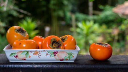 Group of organic persimmons (caqui) fruit whole  in ceramic bowl on wood against tropical backgroundの写真素材