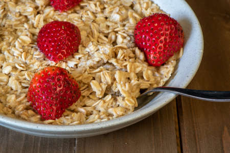 Oatmeal in a tan plate decorated with halved strawberries on wooden table, croped plate, centered, angle view, with silverware spoonの写真素材