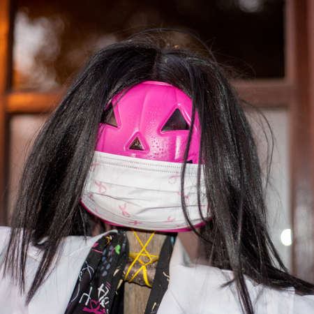 A pink jack-o-lantern scarecrow with black wig wearing a surgical mask decorated with pink ribbon...