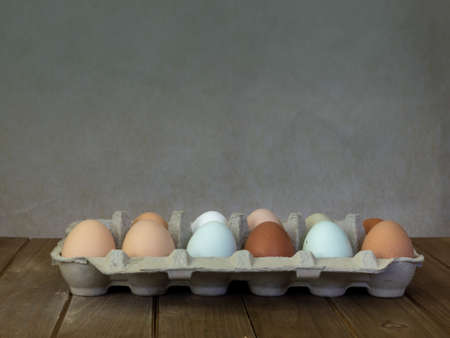 One dozen unwashed colorful chicken eggs in open cartonon on wooden surface viewed from the side and plenty of copy-space- free-range eggs of various shapes, colors and sizes very different from commercial eggsの写真素材