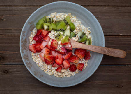 Oatmeal in a tan plate decorated with diced strawberries and kiwi on wooden table, whole plate, centered, top view, with silverware spoonの写真素材