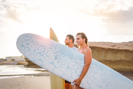 Young couple of happy surfers standing on the beach holding surfboards preparing to surf on high waves during a magnificent sunset - People, lifestyle, extreme sport concept - Focus on manの写真素材