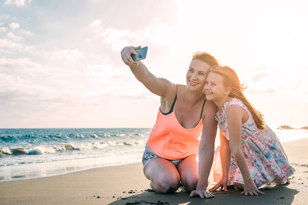 Happy loving family mother and daughter taking a selfie with mobile smart phone on the beach at sunset - Mom with her kid in holiday - Parent, vacation, tech and family lifestyle conceptの写真素材