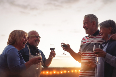 Happy senior friends drinking red wine at barbecue dinner in terrace - Mature people cheering and laughing together at sunset on rooftop - Friendship and elderly retirement lifestyle conceptの写真素材