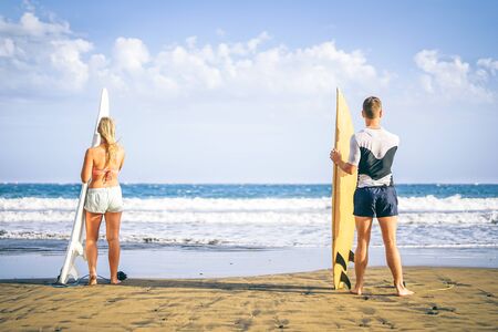 Young couple of surfers standing on the beach with surfboards preparing to surf on high waves