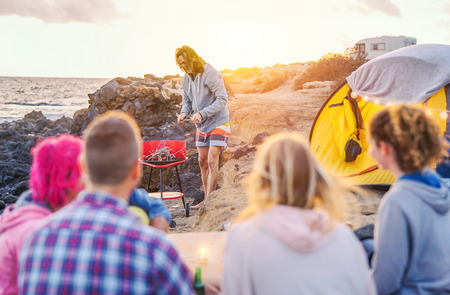 Group of happy friends camping with tent on the beach and preparing a barbecue dinner - Young people...