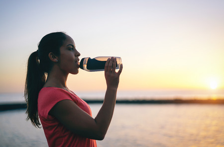 Young sporty woman drinking water while having a break - Health girl resting at sunset after running...