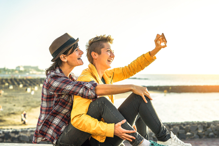 Happy gay couple taking a selfie with mobile smart phone camera on the beach at sunset - Lesbians having fun in holiday - Homosexuality, diversity, vacation, travel, lgbt, technology conceptの写真素材
