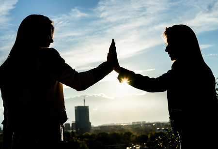Silhouette of two young women giving high five having happy feelings standing on a hill at sunset in a park outdoor with city in the background - Concept of friendship, lifestyle, unity and travelの写真素材