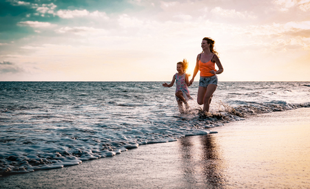 Mother and daughter running along the seashore at sunset - Happy family having fun playing together in vacation on the beach - Parenthood, happiness and love conceptの写真素材