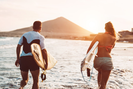 Happy surfer couple running with surfboards along the sea shore - Sporty people having fun going to surf together at sunset - Extreme surfing sport and youth relationship lifestyle conceptの写真素材