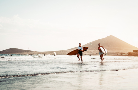 Happy surfer couple running with surfboards along the sea shore - Sporty people having fun going to surf together at sunset - Extreme surfing sport and youth relationship lifestyle conceptの写真素材