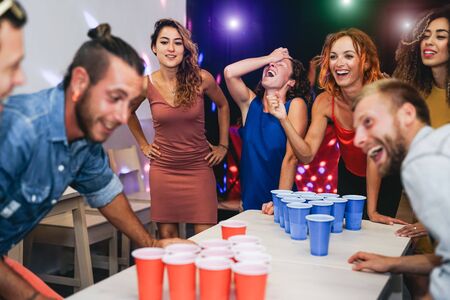 Happy friends playing beer pong in a cocktail bar - Young millennial people having fun doing party alcohol games at night pub - Friendship and youth lifestyle nightlife conceptの写真素材