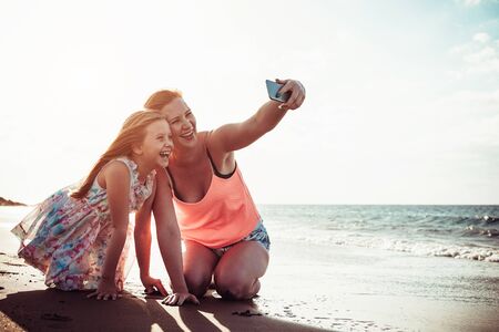 Mother and daughter taking selfie with mobile smartphone while playing on tropical beach at sunset - Happy family having fun with new technology apps for social media - Parenthood conceptの写真素材