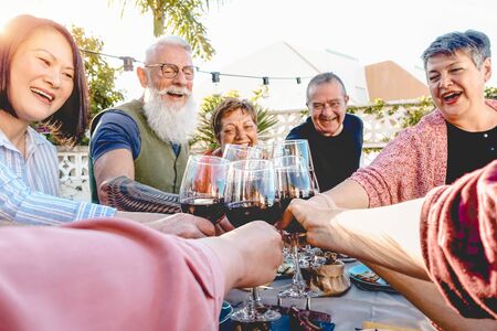 Happy senior friends toasting with red wine glasses at dinner on patio - Mature people having fun dining together outside - Elderly lifestyle, food and drink, retired and pensioners conceptの写真素材
