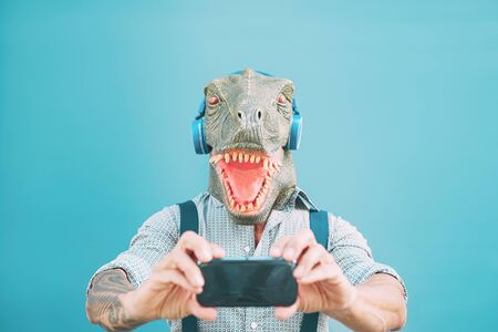 Senior fashion man taking selfie with mobile smartphone while wearing t-rex mask - Hipster guy having fun listening music masquerade with headphones - Funny and absurd surreal conceptsの写真素材