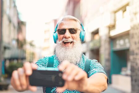 Senior bearded man taking selfie with mobile phone while listening favorite playlist with headphones...
