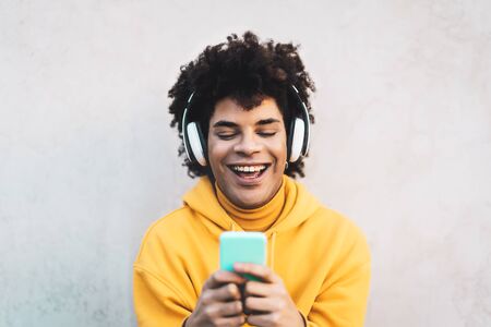 Happy Afro man using mobile smartphone outdoor - Young guy having fun listening music with wireless headphones outdoor - Youth millennial generation lifestyle and people addicted technology conceptの写真素材