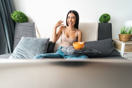 Happy woman eating cereal cup with milk while watching on computer laptop sitting on sofa - Health...