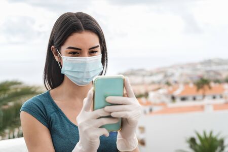 Young woman wearing surgical face mask using mobile smartphone on home terrace - Girl in quarantine having fun with new technology social media apps - Corona virus confinement and youth people conceptの写真素材