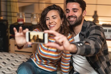 Happy couple taking selfie with mobile smartphone in living room at home - Young lovers having fun with new trendy social media apps - Love relationship technology conceptの写真素材