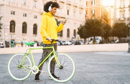Happy Afro man using mobile smartphone outdoor - Young guy having fun listening music with headphones while riding with bike in city - Youth millennial generation lifestyle and technology conceptの写真素材